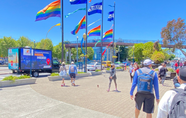 Community vaccination ad campaign using Can't Miss US' mobile billboard parked in a sunny plaza with pedestrians and rainbow flags.