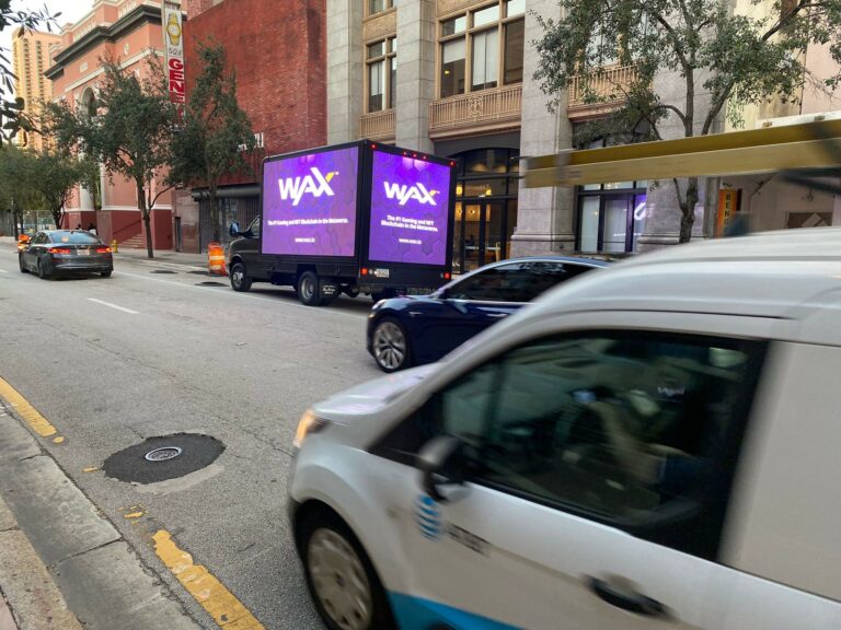 Billboard truck parked in a street with many cars driving by and seeing the WAX ad. Mobile LED billboard shows a blue screen with WAX written