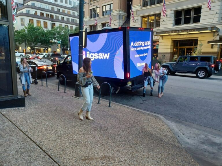 New York digital mobile billboard truck shows Jigsaw Dating App. A woman walking smiles as 2 ladies pose in front of billboard truck.