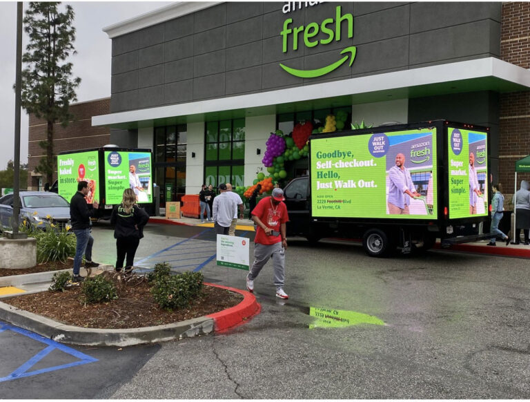 A Cantmiss.US digital billboard truck parked outside an Amazon Fresh storefront on a cloudy day. The truck displays vibrant ads promoting Amazon Fresh's 'Just Walk Out' feature with the message 'Goodbye, Self-checkout. Hello, Just Walk Out.' A depiction of a man on the ad is seen happily exiting after his shopping. Around the store entrance, promotional signs emphasize fresh fruits and vegetables. A steady stream of traffic passes by, and onlookers, including a few who pause to take in the truck's message, navigate the parking lot.