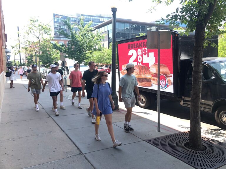 Sunlit city sidewalk with Cantmiss.US digital billboard truck showcasing Wendy's breakfast 2 for $4 offer amid modern buildings and green trees. Pedestrians in summer attire pass by noticing the iconic Wendy’s logo.