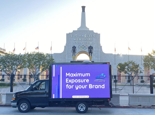 A Cantmiss.US digital billboard truck parked in front of the iconic Los Angeles Memorial Coliseum, illuminated with the message 'Maximum Exposure for your Brand' against a purple backdrop, showcasing the website 'www.cantmiss.us'. The historic coliseum facade, complete with Olympic rings and flying American flags, stands tall in the background.