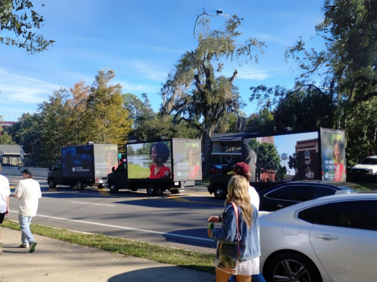 Multiple CantMiss.us mobile billboards parked on a sunny street, prominently displaying advertisements featuring individuals with the captions 'I'm Here Thomasville' and 'Your Neighborhood.' Pedestrians, including a woman in a denim jacket holding a drink, pass by, with trees, cityscape.