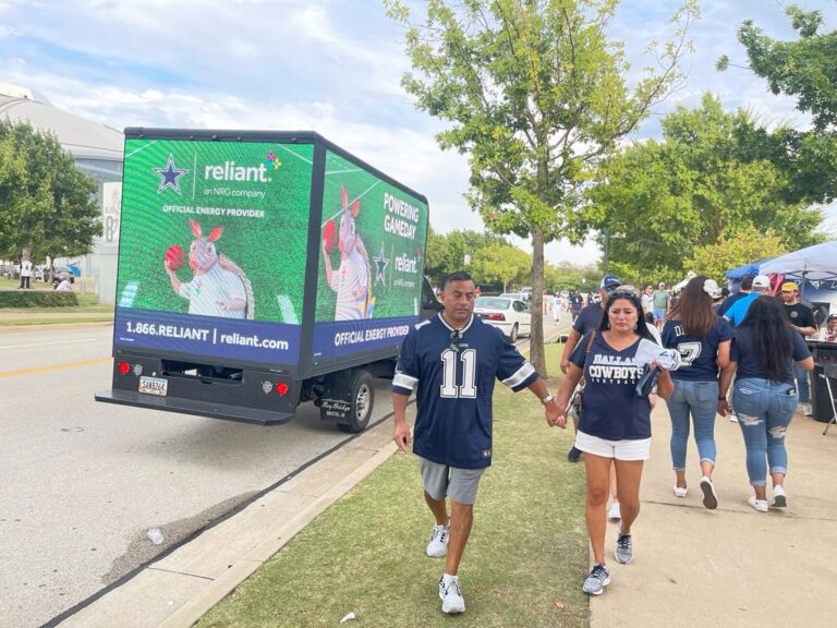 A Cantmiss.US digital LED billboard truck showcasing a vibrant advertisement from Reliant, an NRG company, stands prominently on a street near a park. The display promotes Reliant as the 'Official Energy Provider' with dynamic baseball imagery. Dallas Cowboys fans, wearing their team jerseys, pass by, adding to the game day atmosphere.
