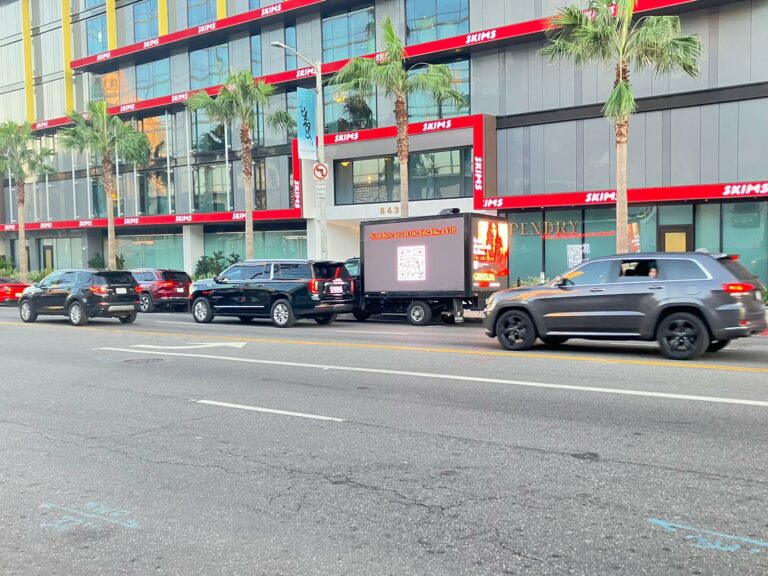Mobile billboard truck parked in between cars on street in front of Store with Skims displaying QR code for Netflix’s Grizelda. Cars drive by