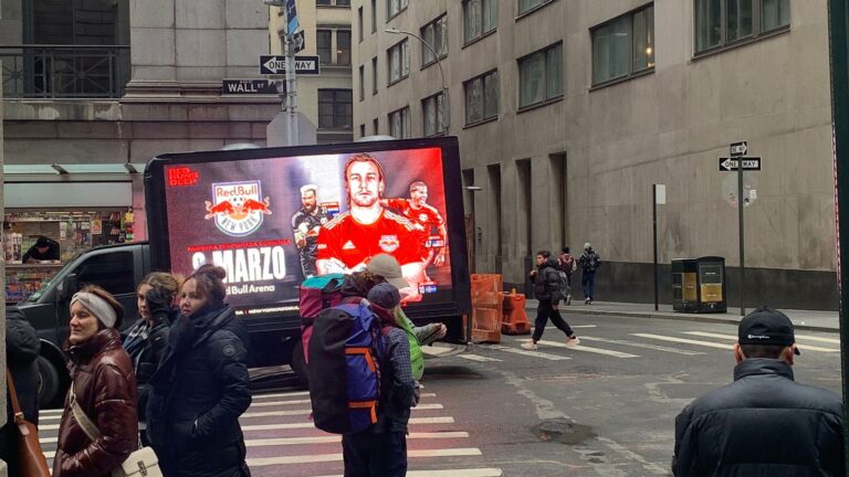 Red bull promotes soccer on mobile billboard. People look at the LED truck with Soccer player wearing red jersey standing with arms crossed with rival team behind him