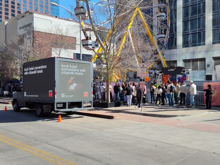 Led advertising truck parked in front of yellow ferriswheel and large number of people. Orange cones front of billboard truck
