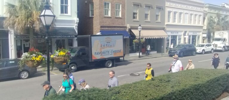 Truck billboard advertising with ad with favorite cheese written in blue parked in front of local shops bldg. Across street, people walking