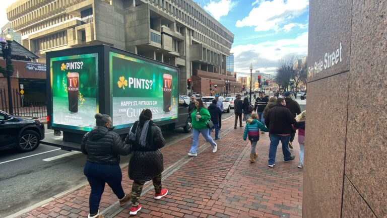 Busy street with billboard advertising truck. Large glass with beer and PINTS? Written. People walking. Woman in green walks close to truck