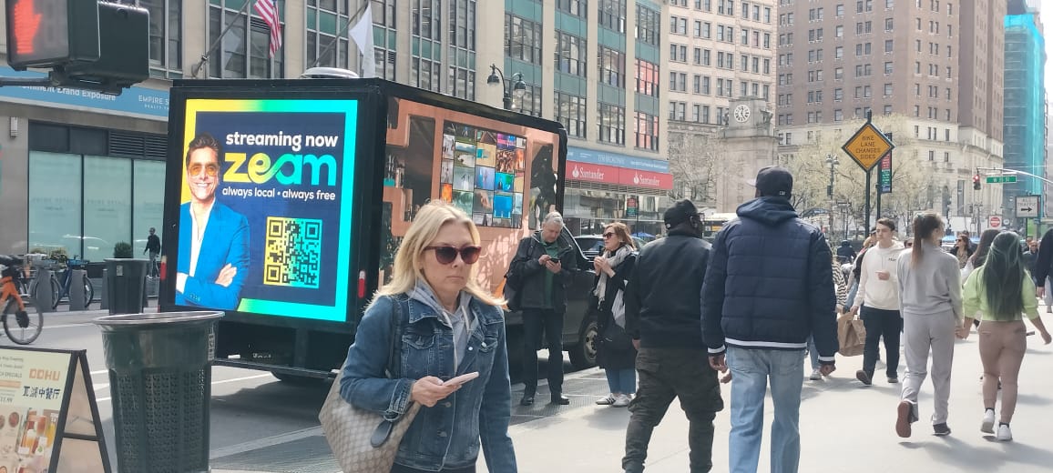 QR code on truck billboard advertising for Zeam streaming.One side of LED shows man with glasses, other side shows a home. Walkers on street