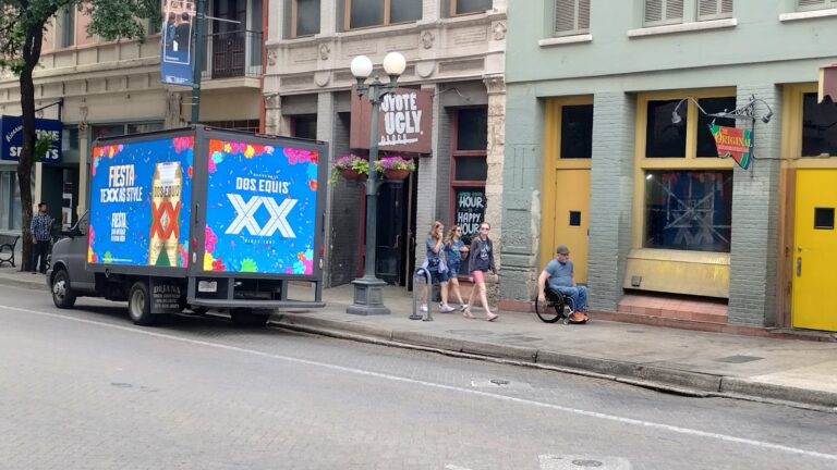 Colorful Dos Equis Mobile Billboard Advertisement truck parked on an urban street with a crowd of people on the sidewalk passing by - Billboard Truck Features