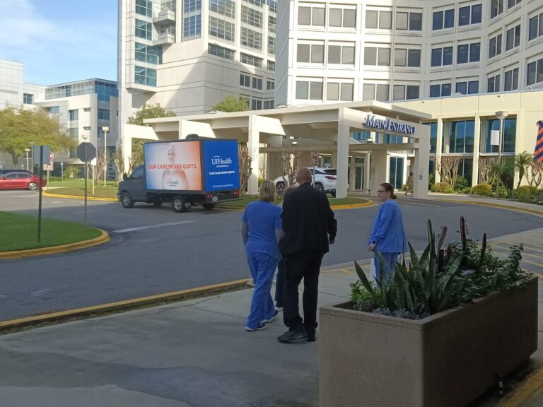 Billboards on trucks with ad for UF Health branding parked beside the hospital main entrance and in front of a sidewalk with a crowd.