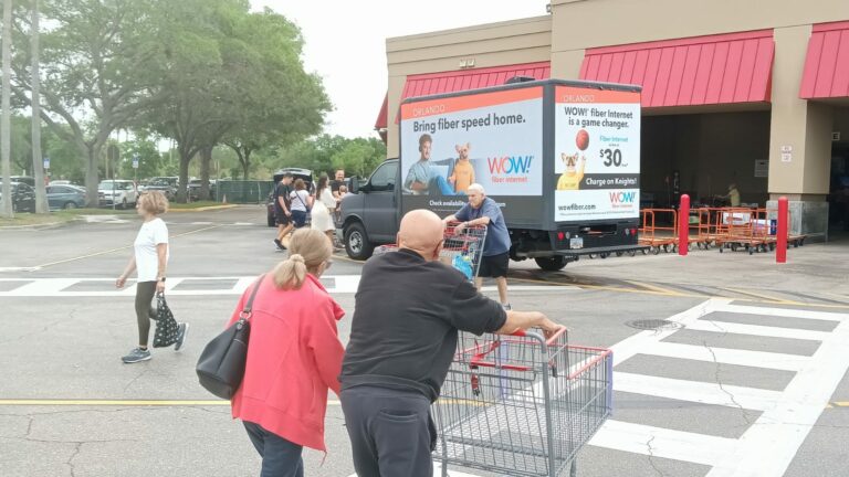 WOW! fiber internet advertisement truck parked beside Orlando shopping center in front of a busy parking area with shoppers passing by.