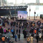 Crowd attends peace event by a historic ship at the harbor, with stage and banners and an LED Mobile truck promoting the event by the corner.