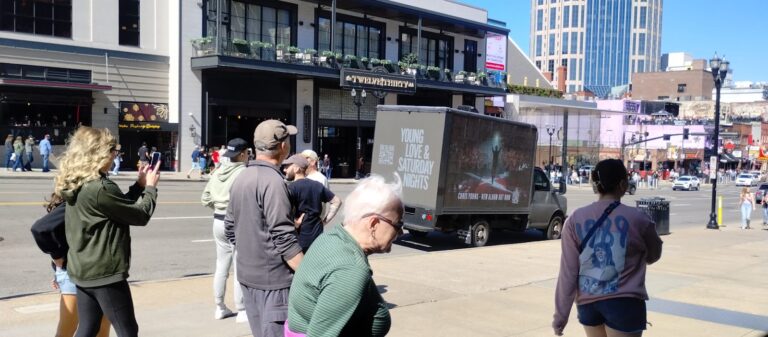People on a city street with LED mobile truck advertising a concert with a message "Young Love and Saturday Nights" on downtown location.