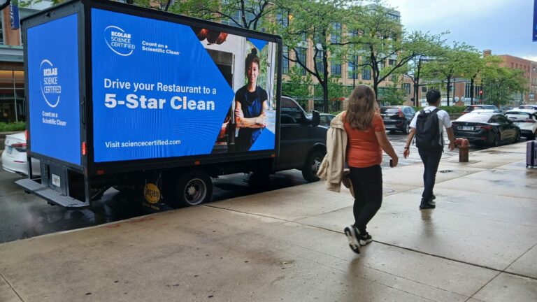 Ecolab Science Certified advertisement on a digital mobile billboard truck parked beside the city street. Several people are walking by.