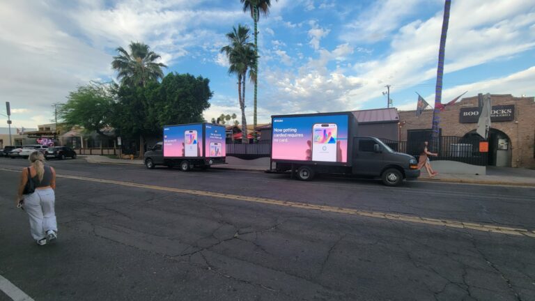 A fleet of mobile billboard trucks promoting Apple Wallet parked in front of a restaurant with several pedestrians passing by.