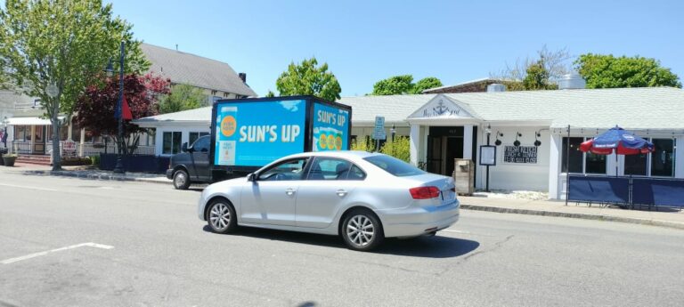 Car driving past a commercial truck with a High Noon drink advertisement with a tagline Suns up in Cape Cod in a Sunny weather.