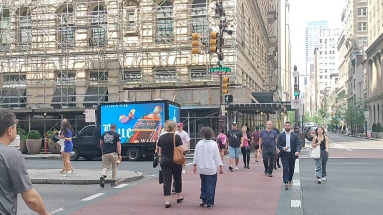 Pedestrians crossing on Broad Street in Philadelphia with a parked Digital Mobile Billboard Advertisement truck for Johnnie Walker.