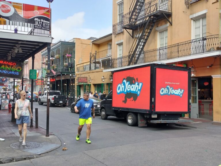 Digital billboard truck advertisement for Kool-Aid parked on a busy sidewalk in front of a local shop on a vibrant New Orleans with several people walking by.