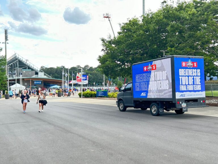 Commercial truck with an ACC advertisement parked near a sports stadium with people walking in the area on a fine weather.
