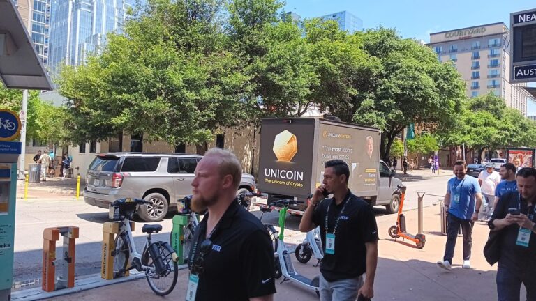 Several people are walking past a bicycle rack and a LED advertising truck for Unicoin parked on a street in Austin, Texas.