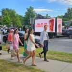People walking on a sidewalk next to a street in a city with a Dunkin Donut advertisement on a mobile digital billboard truck.