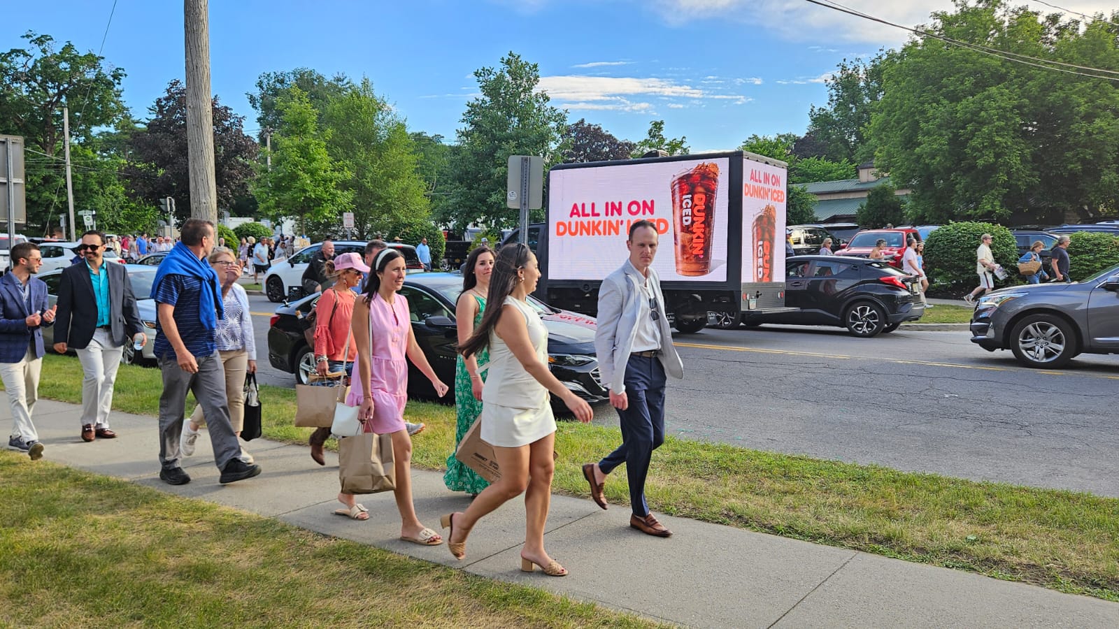 People walking on a sidewalk next to a street in a city with a Dunkin Donut advertisement on a mobile digital billboard truck.