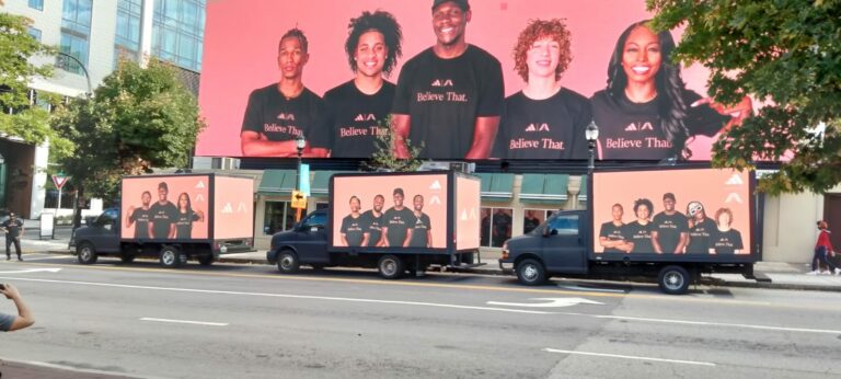 A fleet of outdoor advertising trucks with 'Believe That' promotion parked beside a sidewalk in Atlanta, promoting diversity.