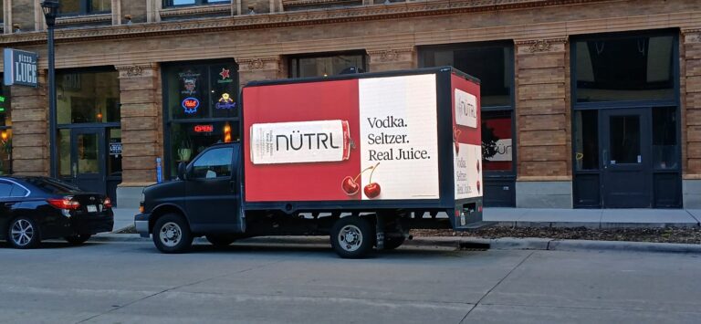 NÜTRL advertisement with the words "Vodka. Seltzer. Real Juice." on a mobile digital billboard truck parked in front of a building in a city street.