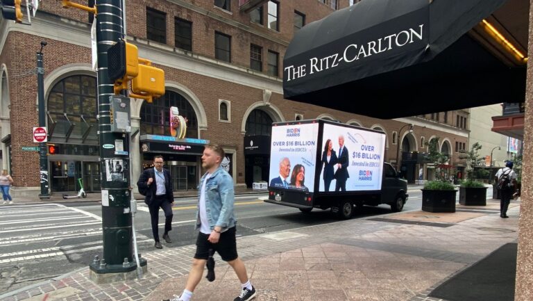 People walking past an outdoor advertising truck for Biden-Harris parked on the side of the road near The Ritz-Carlton, downtown.