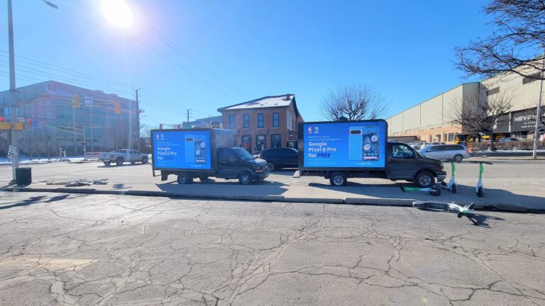 A fleet of LED advertising trucks for Google Pixel 8 Pro promotion with a slogan "Elevate your game" is parked on a city street in a sunny weather.
